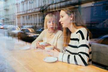View through the window of young beautiful blonde girl talking to her mom an interesting secret, they sit at cafe, happy and laugh. Bonding, different generationsconnect, family concept.