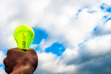 Green bulb in hand on blue sky background. Hand holding light bulb on a background sky and sun. The energy of the sun and the wind, the concept of pure enegry