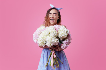 Cheerful young lady with long brown hair  being excited to get bouquet of spring flowers on women's day isolated over pink background.
