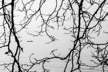 Fototapeta na wymiar Black and white photo. View of the silhouette of a boat with fishermen through the tree branch. Silhouette photo. Moody landscape. Lake Garda, Italy