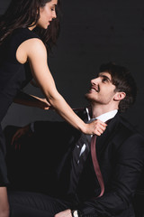 attractive brunette woman in black dress taking off tie with handsome man in suit sitting on black background