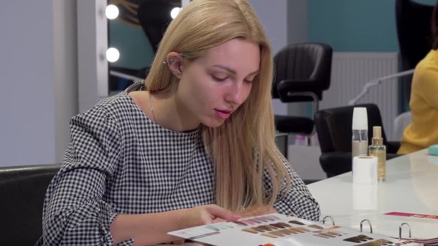 Young woman choosing new hairstyle from catalogue at hairdresser salon. Blond female customer waiting for her stylist at beauty studio. Beauty, hair care concept
