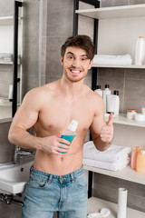 handsome shirtless man smiling, holding mouthwash and showing thumbs up with hand in bathroom