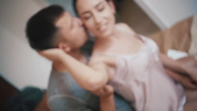 Young happy couple in love having fun together in bedroom at home, playful wife biting smiling husband ear, embracing, piggyback, man and woman playing childish in bed, enjoying funny intimate moments