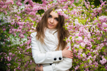 A beautiful girl of Slavic appearance in a white coat stands in the Japanese Sakura garden and hugged herself. Cherry blossom