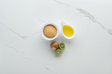 top view of whole and cut kiwi, bowls with brown sugar and oil on white surface