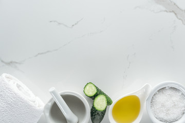 top view of bowls with oil and salt, cucumber halves, pounder and towel on white surface