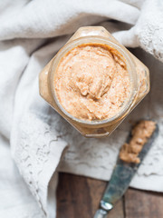 Vertical photo of homemade natural almond butter in a glass jar placed on rustic beige table cloth and with a butter knife on the side. Pastel colors. View from above. 