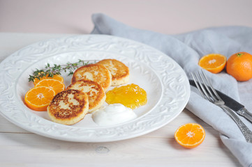 Cheesecakes with citrus jam and sour cream on a white plate. Next to the plate is a cloth napkin and cutlery. The composition is complemented by slices of mandarin. Light wooden background. Close-up.