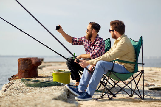 leisure and people concept - friends with fishing rods taking selfie by smartphone on pier at sea