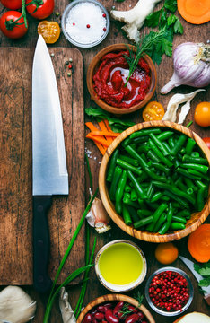 Green beans with roots, vegetables, mushrooms, spices and tomatoes, vegan bowls. Food cooking background, vintage wooden rustic table. Big chef's knife, vertical image. View from above