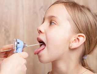 Pediatrician examining elementary age girl sore throat using wooden tongue depressor and torch in...