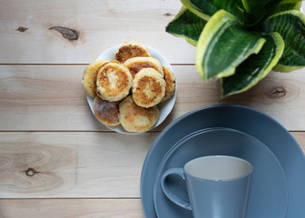 cottage cheese pancakes on a saucer on a wooden surface, next to a stack of clean blue plates, green flower in a pot, top view