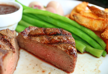 Closed up medium grilled tenderloin steak cut in half with blurry steamed vegetables and fried Potatoes served on white plate