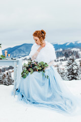 Fototapeta na wymiar Attractive red-haired girl with a bouquet in her hands sitting on a chair against the backdrop of winter mountains