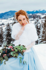 Stunning red-haired girl with a bouquet in her hands sitting on a chair against the backdrop of winter mountains