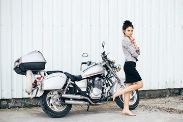 Fototapeta na wymiar Young pretty cute brunette girl dressed in a little black dress and a jacket standing near a motorcycle