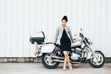 Obraz na płótnie Canvas Young pretty cute brunette girl dressed in a little black dress and a jacket standing near a motorcycle