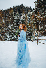 A charming red-haired girl in a blue dress standing in a snowy lawn with a bouquet in her hands