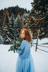 A charming red-haired girl in a blue dress standing in a snowy lawn with a bouquet in her hands