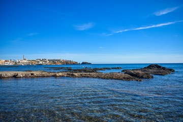 Crystal clear water envelop dark grey rocks that just rise above the surface, with the small village of Cabo de Palos, Spain in the background.