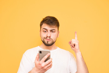 Emotional man with a beard holds a smartphone in his hands, showing his finger up in a blank space. Surprised adult in a white T-shirt uses a smartphone and shows a finger on copyspace