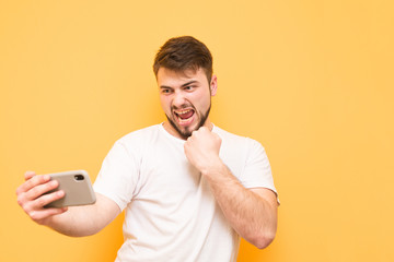 Man with a beard plays a video game on a smartphone, wears a white T-shirt, looks at the phone screen and rejoices in victory. Winner with a smartphone in his hands is isolated on a yellow background.