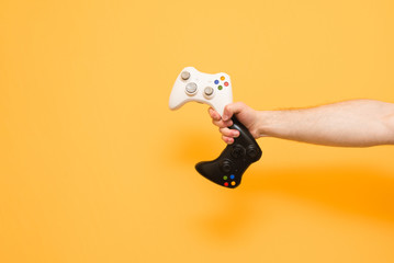 Man holds two joysticks in his hand, white and black on a yellow background. Hand with the gamepad is isolated on a yellow background.Background. Copyspace. Game Concept.