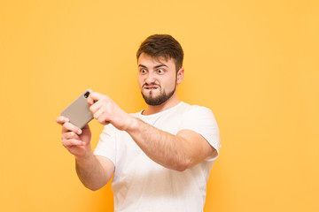 Emotional evil man with a beard playing mobile games on a smartphone, focused on looking at the screen on a yellow background. Adult wears a white T-shirt and plays games on a gadget. Mobile gaming.