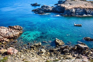 Crystal clear water envelop dark grey rocks that just rise above the surface, with a small boat and some people snorkelling.