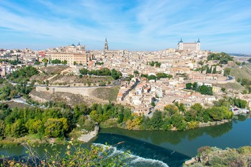 Fototapeta na wymiar A panoramic view of the old city of Toledo, with it’s defensive walls, the tower of the cathedral and the old castle on top a hill inside the city walls, surrounded by a river that serves as a moat.