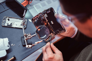Repairman uses magnifier and tweezers to repair damaged smartphone. Close-up photo of a...