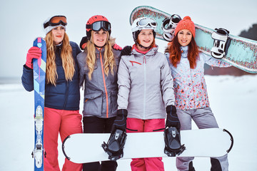 Cheerful women friends in sports winter clothes with snowboards and skis posing and smiling at a...