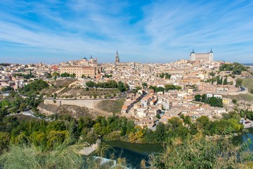 Fototapeta na wymiar A panoramic view of the old city of Toledo, with it’s defensive walls, the tower of the cathedral and the old castle on top a hill inside the city walls, surrounded by a river that serves as a moat.