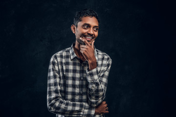 Young dark-skinned guy in shirt smiling and posing standing, looking sideways, putting his hand on a bodbeard in the studio on a black background