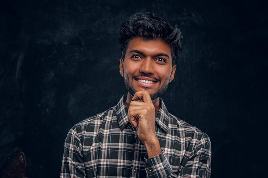 Handsome Indian Guy In Plaid Shirt Smiling And Looks At The Camera With A Piercing Look. Studio Photo Against A Dark Textured Wall