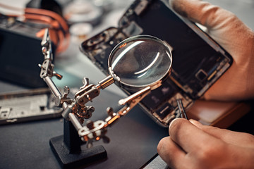 Repairman uses magnifier and tweezers to repair damaged smartphone. Close-up photo of a disassembled smartphone.