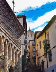 A narrow alleyway in Toledo, Spain going uphill towards a tower pointing to streaks of blue in a cloudy sky
