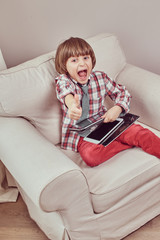 Cheerful young schoolboy wearing a checkered shirt with tie showing trump up and holding a tablet...