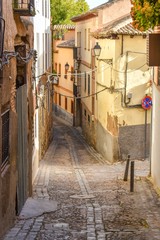 A narrow alleyway in Toledo, Spain, lined with cobblestones and bricks, with cables spanning the way linking tiled roof houses on both sides.