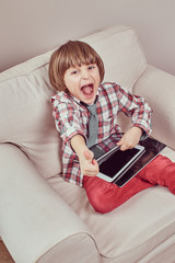 Cheerful young schoolboy wearing a checkered shirt with tie showing trump up and holding a tablet...