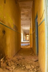 A corridor filled with dirt and broken tiles, with the sun streaming in through the doors of this abandoned building.