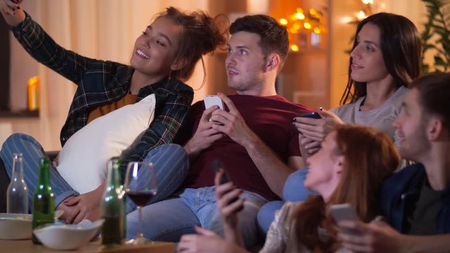 friendship, people, technology and entertainment concept - happy friends with snacks and non-alcoholic drinks watching tv and taking selfie by smartphone at home in evening