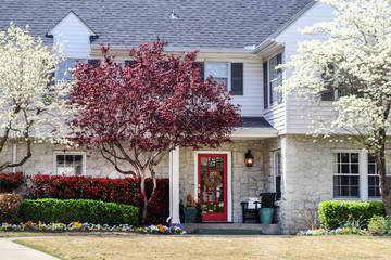 Pretty two story house with rock bottom and white frame with shutters on top in springtime with pansies and flowering trees and wreath on door