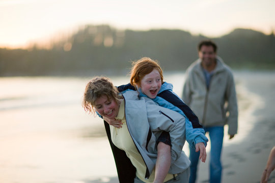 Mid-adult woman piggy-backing her young daughter while walking along a beach at sunset.