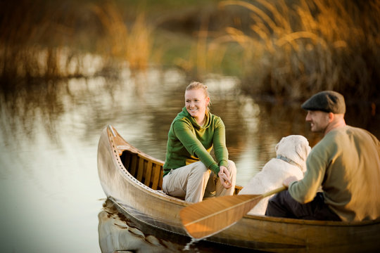 Young adult couple with a dog rowing along a lake in a canoe at sunset.