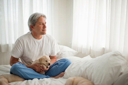 Middle aged man holding Labrador puppy while sitting on his bed.