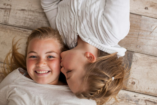 Portrait of a girl being kissed on the cheek by her younger sister while lying on a wooden floor.