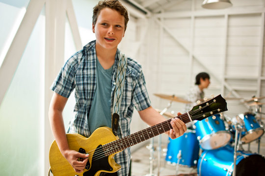 Smiling teenage boy playing an electric guitar while a friend plays the drums inside a garage.