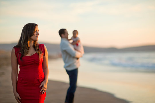 Pensive young pregnant mother looks thoughtfully out to sea while her husband plays with their baby daughter in the background.